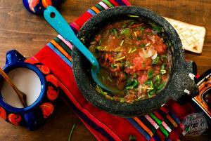 A molcajete filled with chunky salsa molcajeteada sits on a vibrant, striped Mexican cloth. A blue spoon rests inside the bowl. Colorful pottery and a piece of flatbread are nearby on the wooden table.