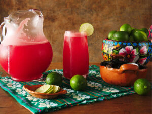 A glass pitcher filled with agua de sandia or watermelon agua fresca, there's also a tall glass with ice cubes filled with it and lime slices.