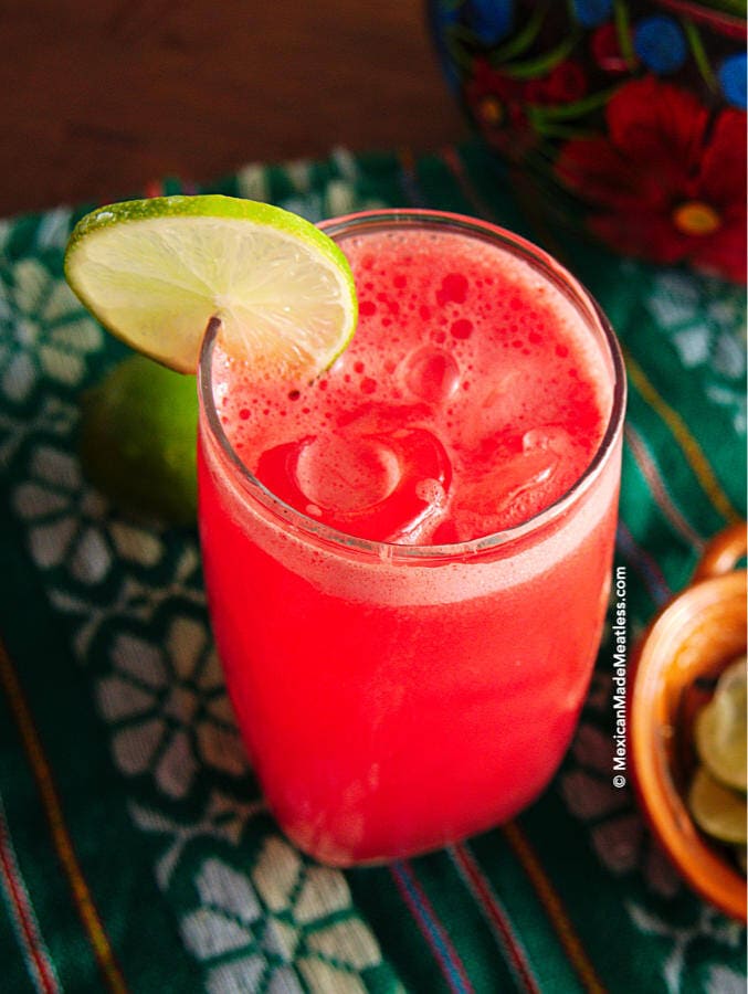 Mexican agua de sandia or watermelon agua fresca inside a tall glass filled with ice cubes and a lime slice on the rim.
