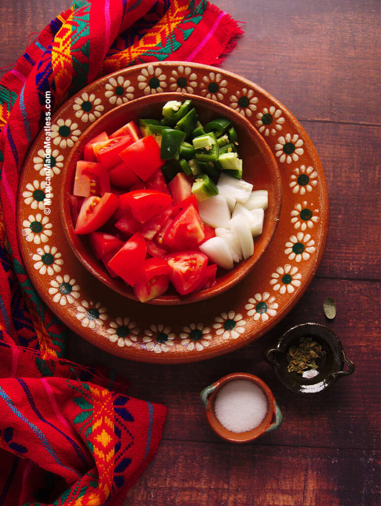 A Mexican teracotta plate with chopped tomato, white onion, jalapeno. On the side are small bowls of salt and dried oregano. These are ingredients for raw tomato salsa.