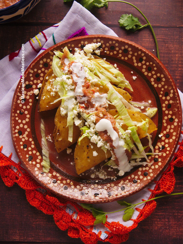 A plate on top of a white and orange kitchen towel, on the plate are 3 crispy fried potato tacos that are topped with lettuce, cheese, salsa and sour cream.