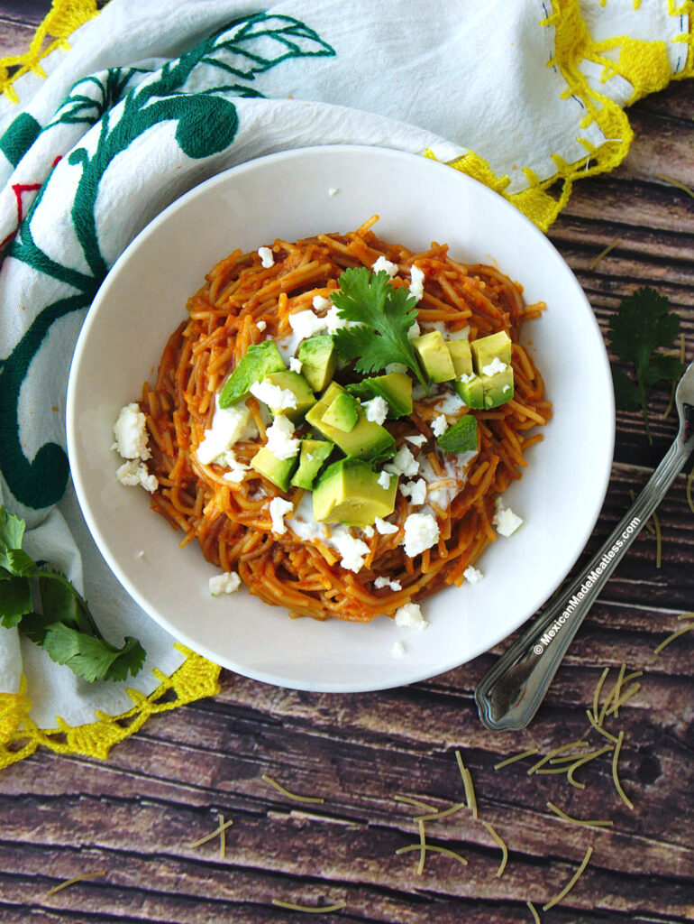 A white bowl of Mexican fideo seco pasta made with tomato sauce and topped with sour cream, cheese and avocado.