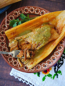 One vegan red tamale on top of a corn husk drizzled with salsa verde.