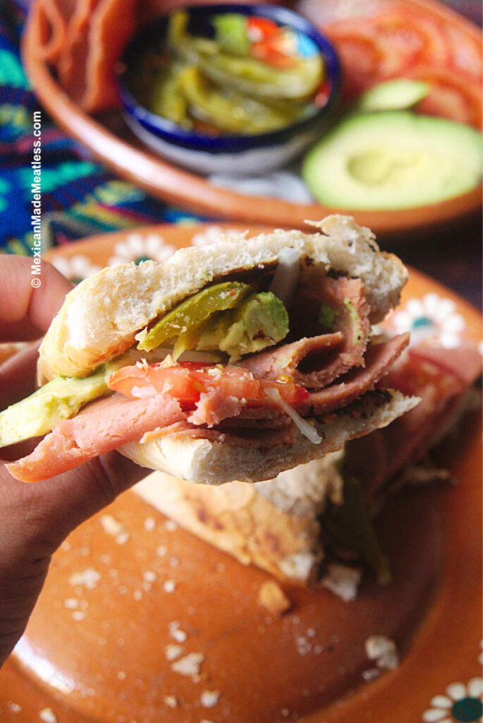 A Mexican torta sandwich made with vegan ham, tomato, onion, avocado and pickled jalapenos. 