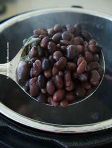 Cooked black beans inside a metal spoon over a pressure cooker.