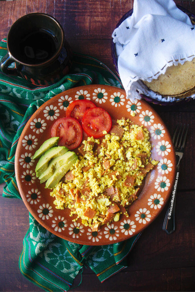 Mexican terracotta plate with vegan ham and eggs, sliced avocado and sliced tomatoes sitting on top of a green cloth on a wood table. There's also a cup of coffee and some corn tortillas inside a basket with a white cloth.