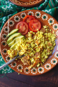 Mexican scrambled ham and eggs made with tofu and vegan ham. There are slices of avocado and tomato on the side.