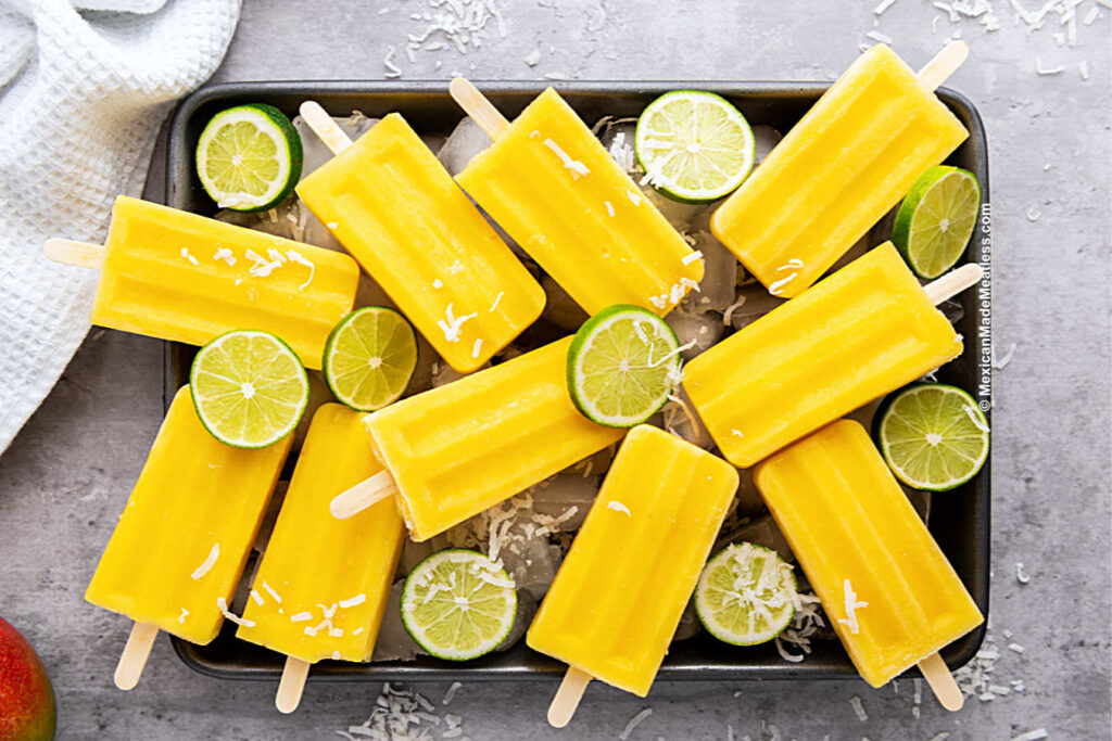 Mexican mango paletas laying on top of ice cubes on a metal tray.