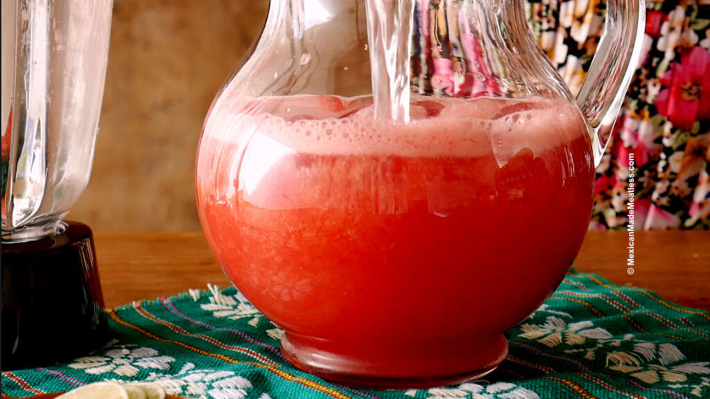 Mixing blended watermelon agua fresca with water inside a glass pitcher. 