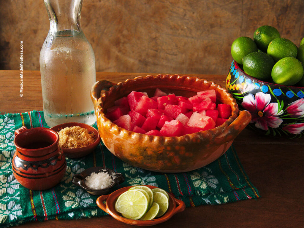 Ingredients for making Mexican watermelon agua fresca inside small bowls. They are watermelon chunks, lime slices, muscovado sugar, lime juice, water and salt.