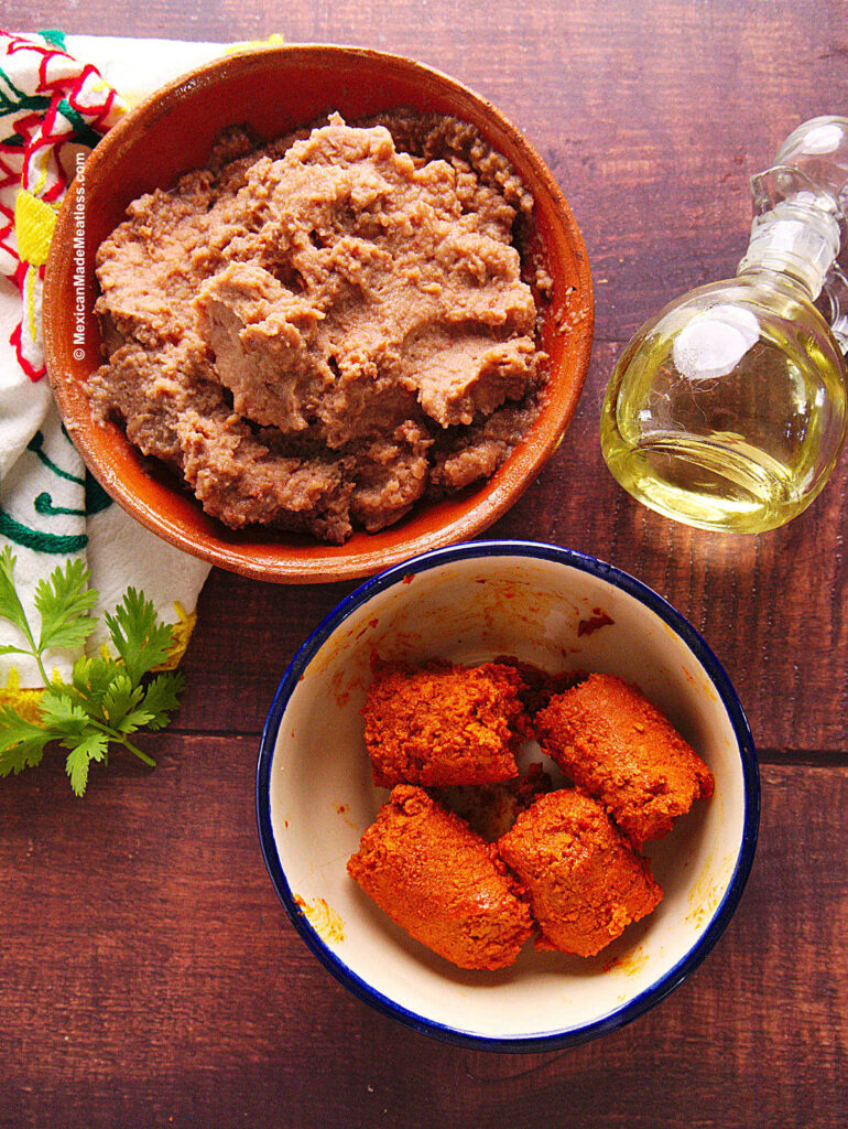 Refried beans in a small bowl, soy chorizo in a small white bowl and olive oil in a glass container. 