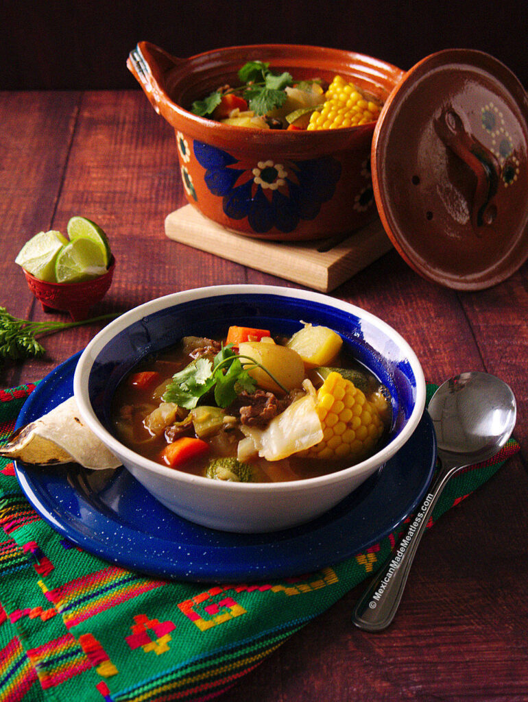 A blue and white bowl filled with Mexican vegan caldo de res or beef stew. There's a corn tortilla and lime wedges on the side.