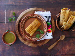 One cooked bean and cheese tamale on a brown plate still wrapped in corn husk.