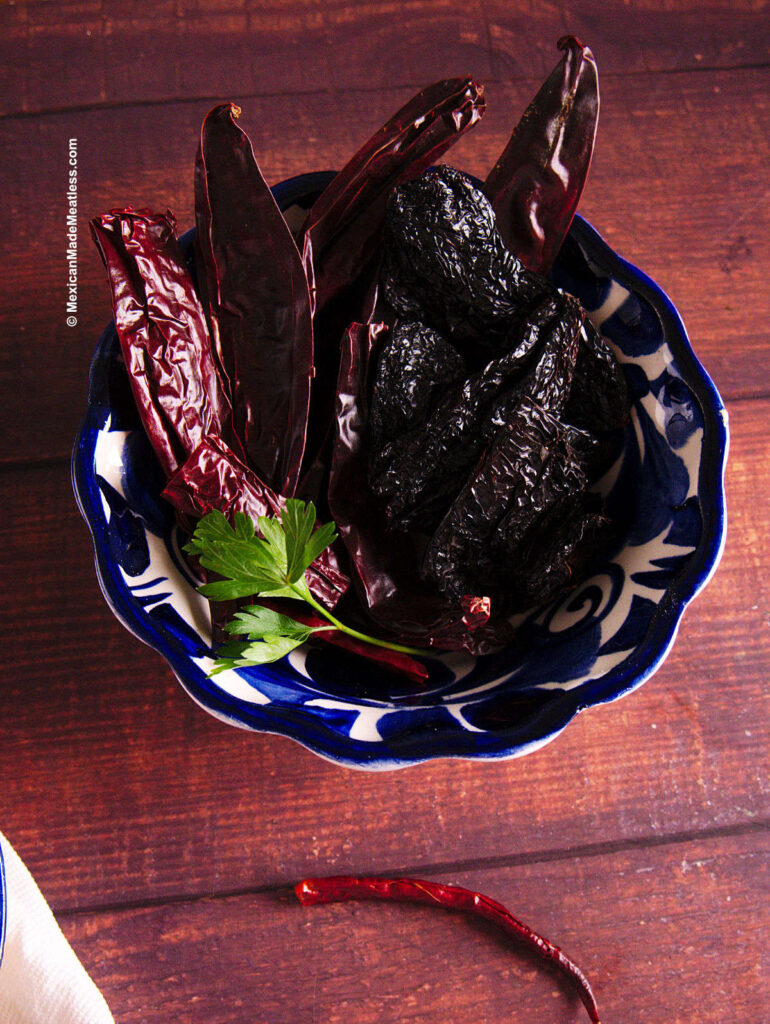 Dried ancho chile, guajillo chilies and chile de arbol inside a blue and white bowl on a brown table.