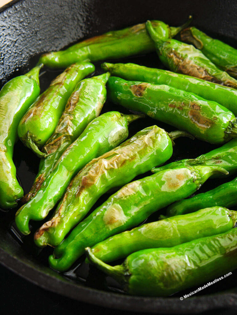 A cast iron skillet filled with small green Japanese peppers called shishitos being fried in a little oil.
