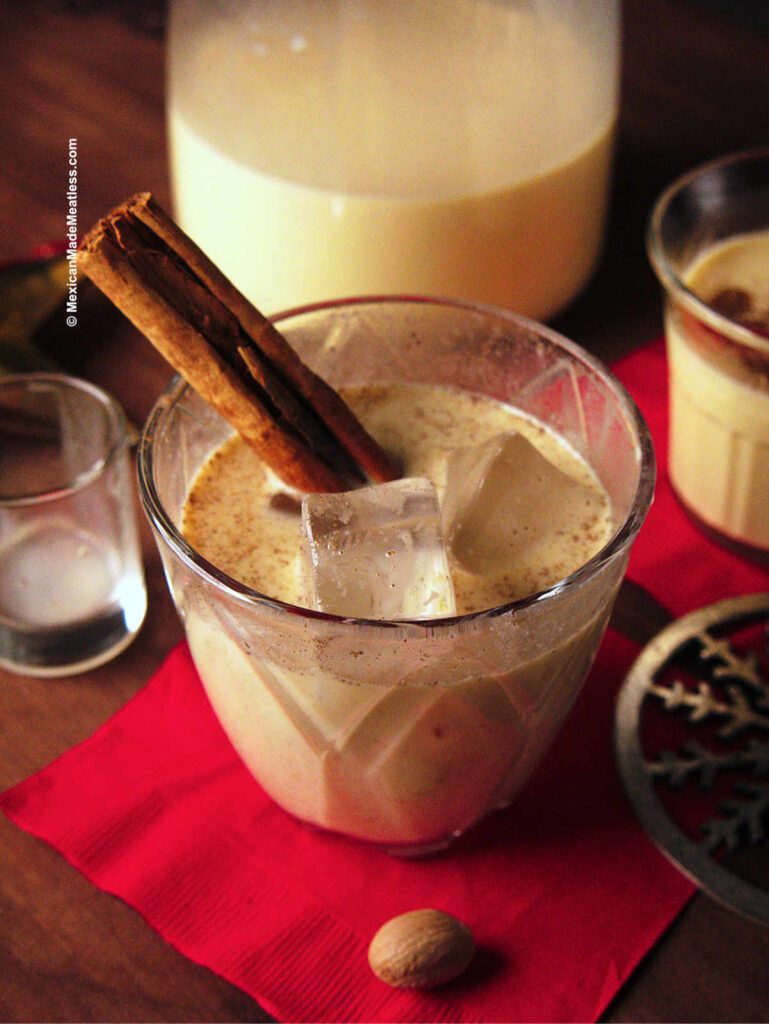 A clear glass full of old fashioned eggnog served over ice and with a cinnamon stick sticking out of it. 