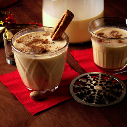 A clear glass full of old fashioned eggnog served over ice and with a cinnamon stick sticking out of it.