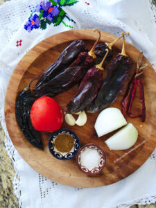 A wooden platter filled with dried Mexican chiles, garlic, onion, roma tomatoes , and spices.