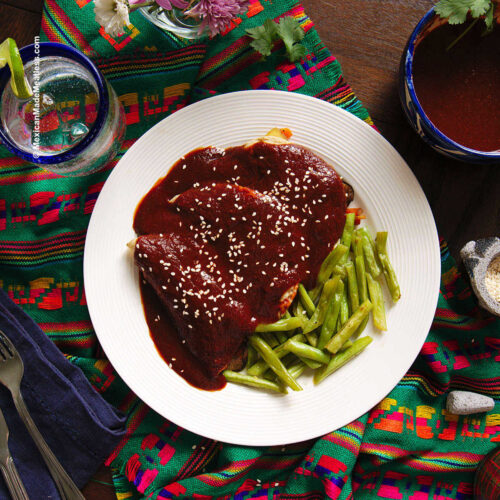 A white plate with three mole enchiladas sprinkled with sesame seeds and some green beans on the side.