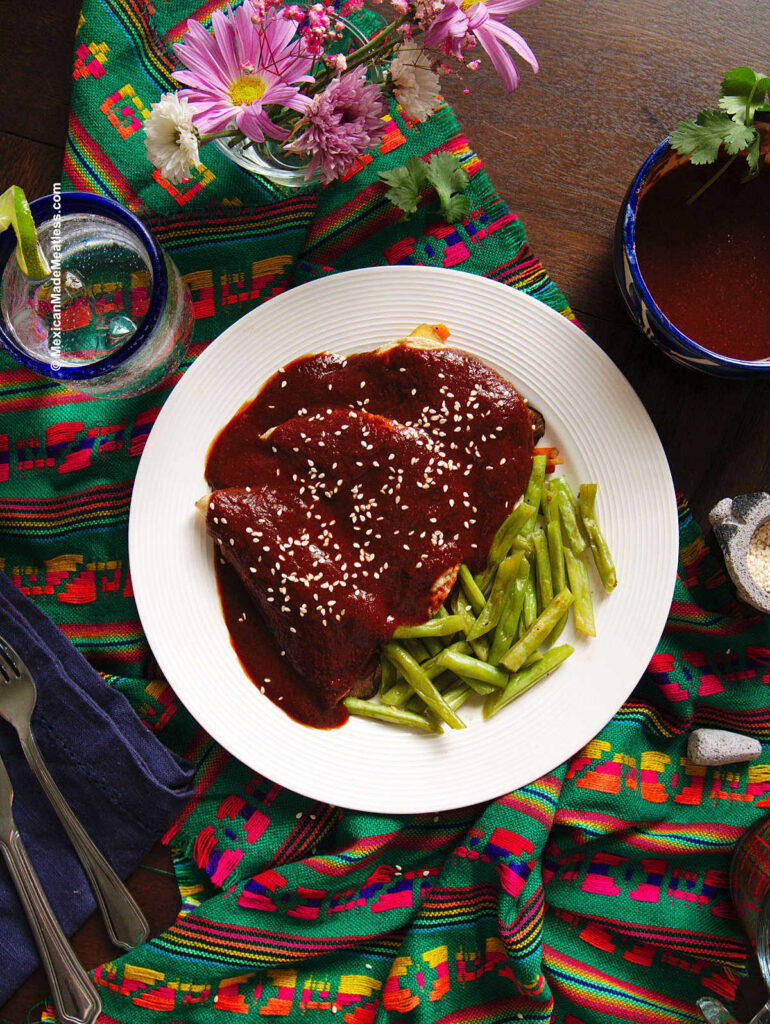 A white plate with three mole enchiladas sprinkled with sesame seeds and some green beans on the side. 