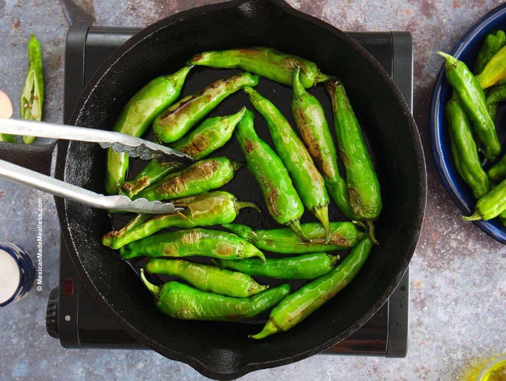 A cast iron skillet filled with small green Japanese peppers called shishitos being fried in a little oil. 