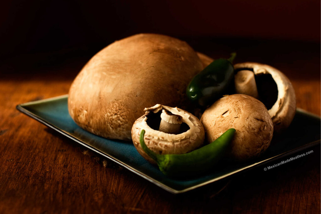 Two portobello mushrooms and 4 small cremini mushrooms with 2 raw jalapeno peppers on a square plate.
