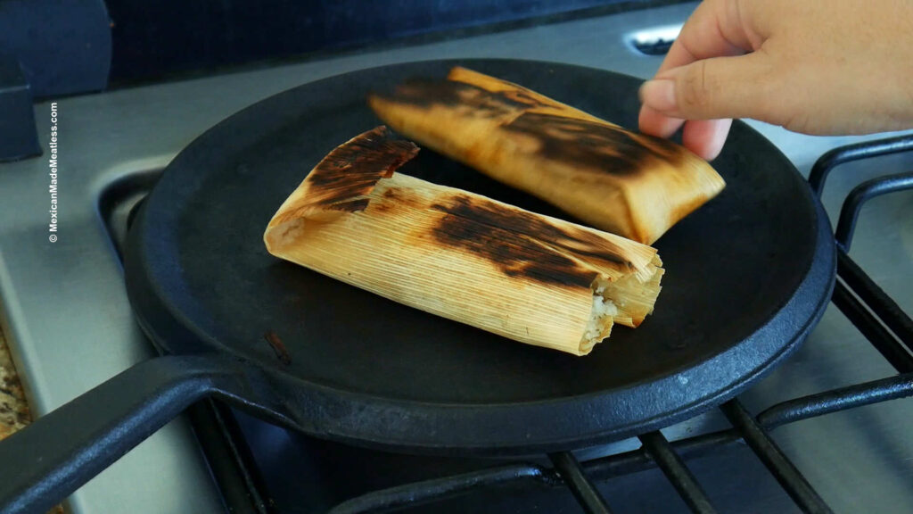Reheating two Mexican tamales wrapped in corn husks on top of a hot cast iron flat grill. 