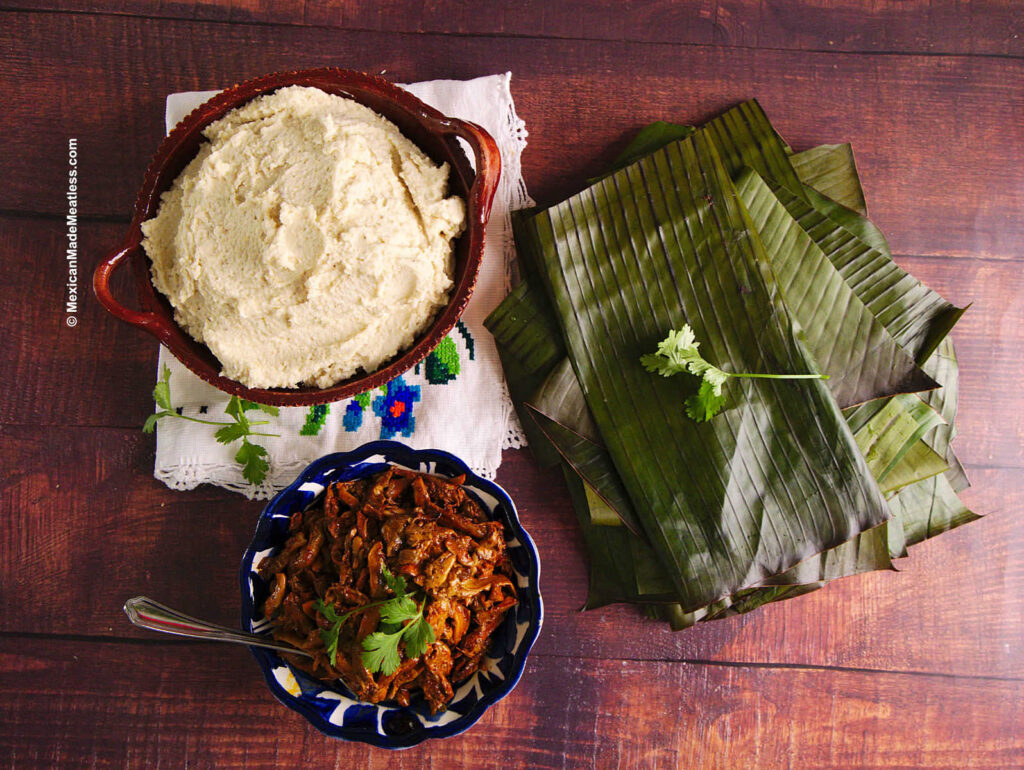 Tamale masa inside a big bowl, fresh banana leaves, and vegan cochinita pibil on top of a brown table. 