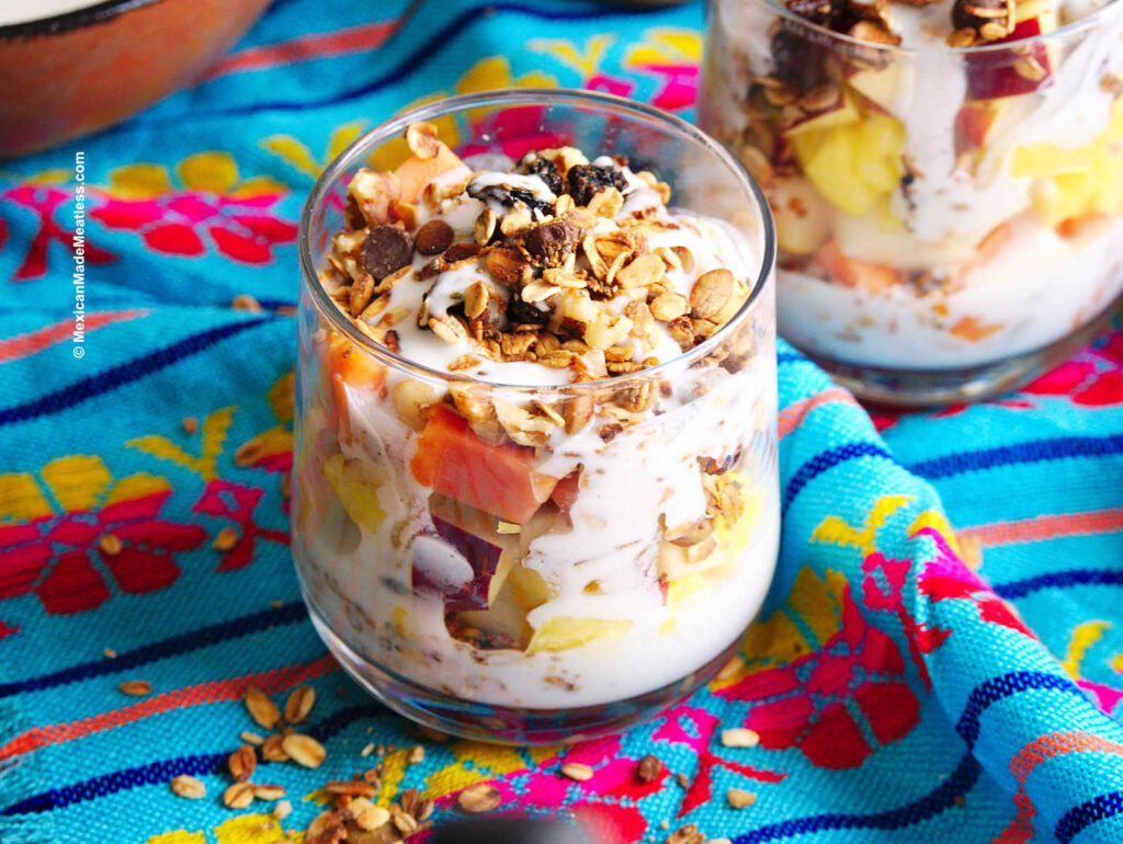 A cup filled with Mexican bionicos fruit salad with sweet cream sauce, granola, nuts and coconut flakes.