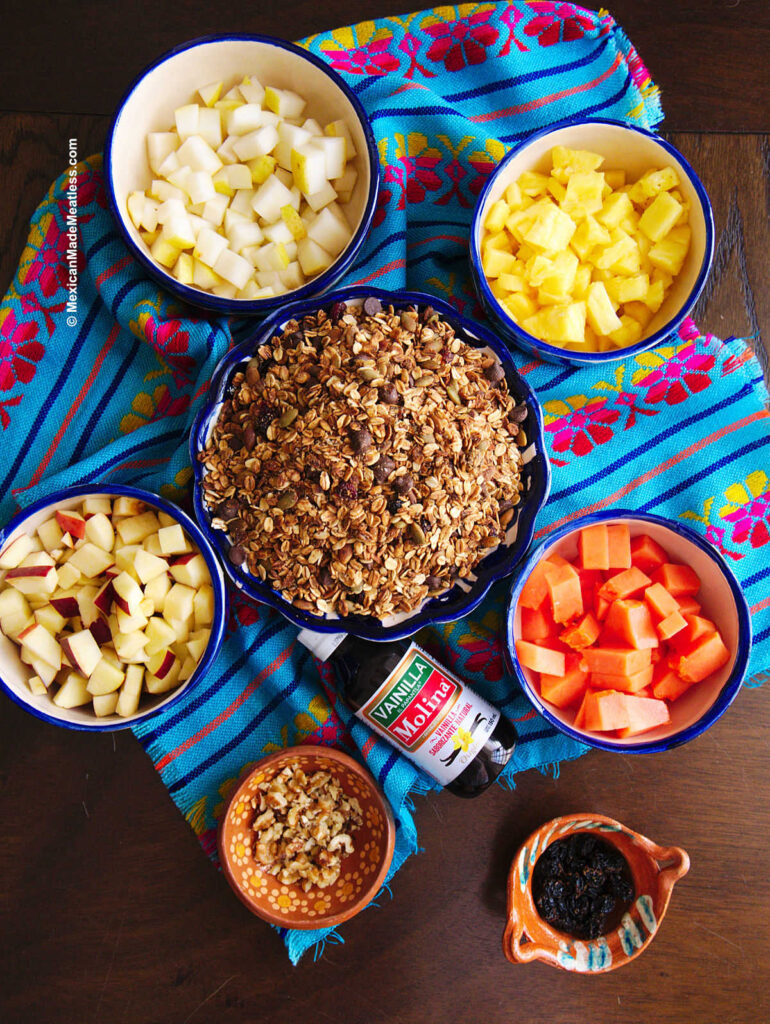 Small bowls filled with fresh chopped fruit, granola, chopped walnuts and raisins on a small brown table. 
