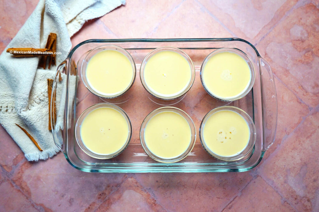 Six small flan glass cups inside a large Pyrex baking pan with  water ready to be baked.