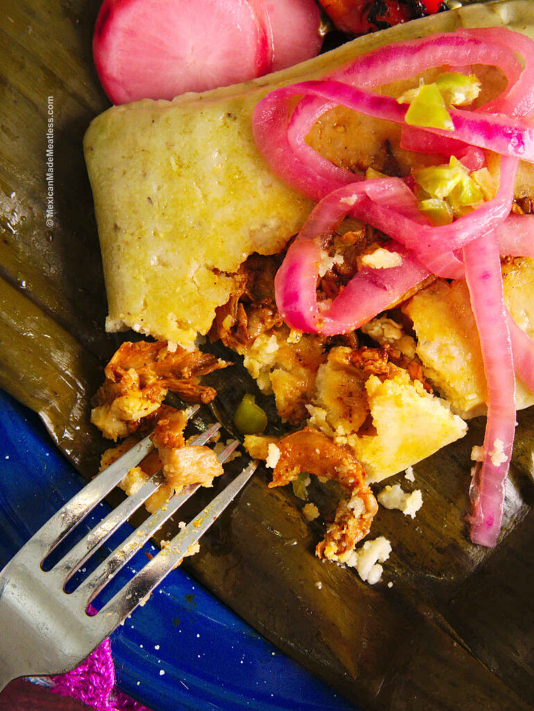 Close up view of a banana leaf tamale stuffed with cochinita and topped with pickled onions.