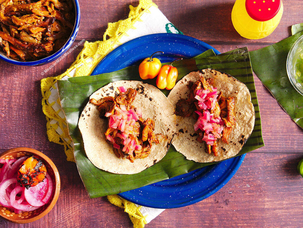 A blue plate with a piece of banana leaf and two cochinita pibil tacos with chopped pickled onion.