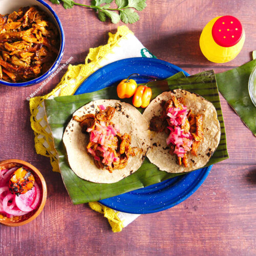 A blue plate with a piece of banana leaf and two cochinita pibil tacos with chopped pickled onion.