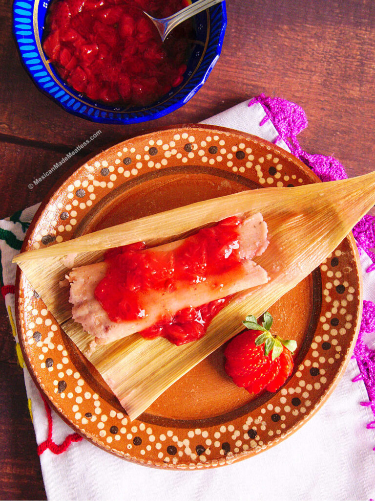 One strawberry tamale drizzled with strawberry jam laying on top of a corn husk that's on a decorative Mexican plate.