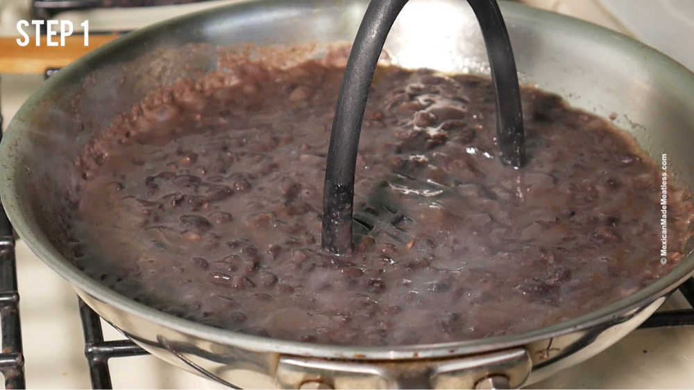 A stainless steal pan with black beans and a potato masher for making refried beans.