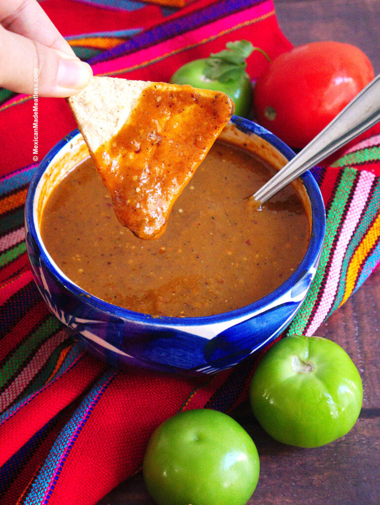 A small blue bowl filled with spicy salsa taquera and a tortilla chip being dipped into it.