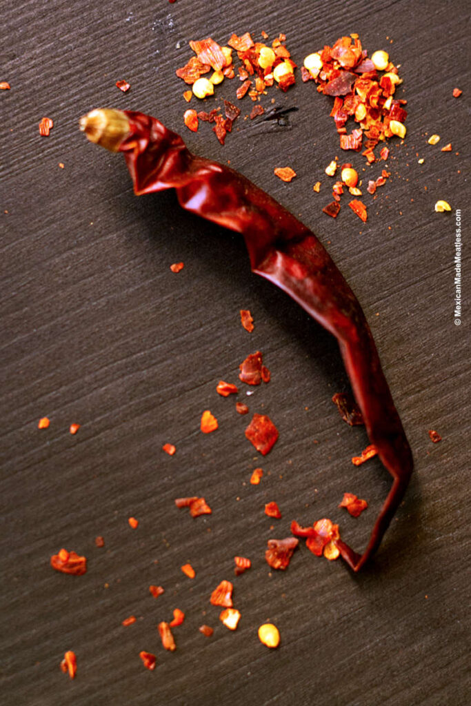 A black table with one whole dried red chile and red pepper flakes.