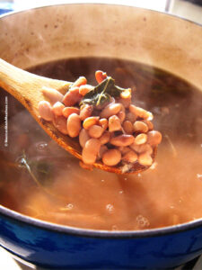A wood spoon scooping out cooked pinto beans from a blue pot.