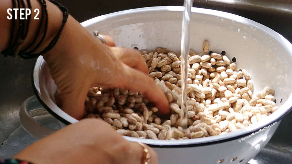 A white colander filled with dried pinto beans being rinsed under cool water in the kitchen sink.