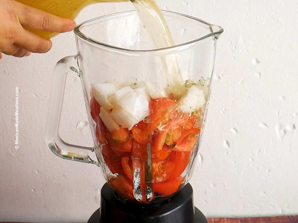 A glass blender cup and a woman's hand placing chopped tomato, onion, garlic, dried chile and vegetable broth into it.