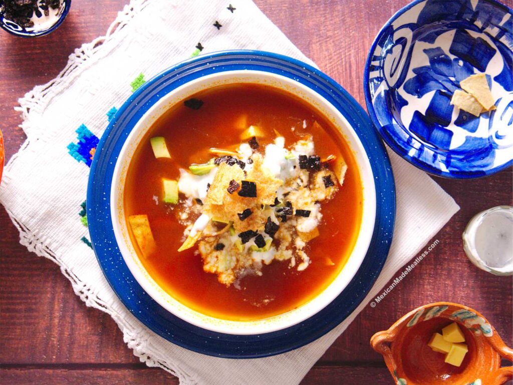 A white bowl on top of a blue plate that's filled with Mexican tortilla soup and garnishes.
