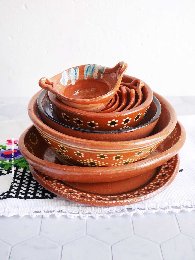 Mexican barro plates and bowls stacked on top a decorative tablecloth.