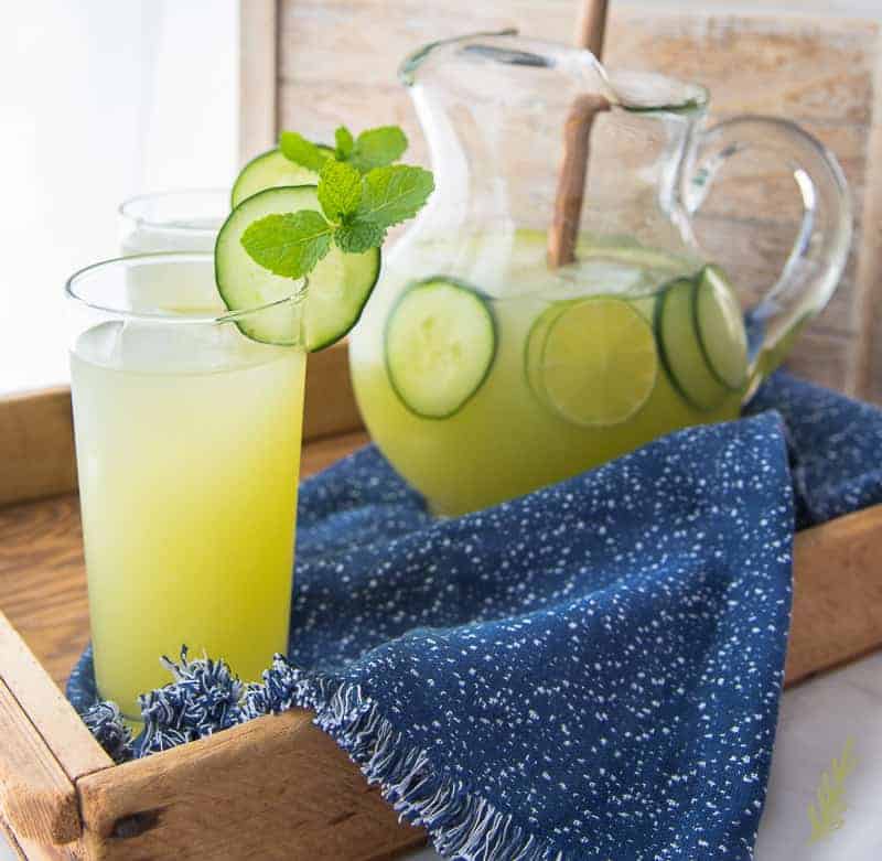 Two tall glasses and one glass pitcher full of cucumber lime agua fresca with cucumber slices.