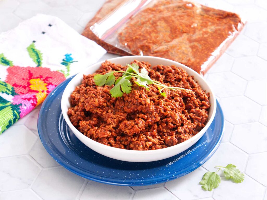 A white bowl placed on top of a blue plate and the bowl is filled with homemade Mexican vegan chorizo made from soy crumbles. Additionally there's two ziploc bags filled with the chorizo.