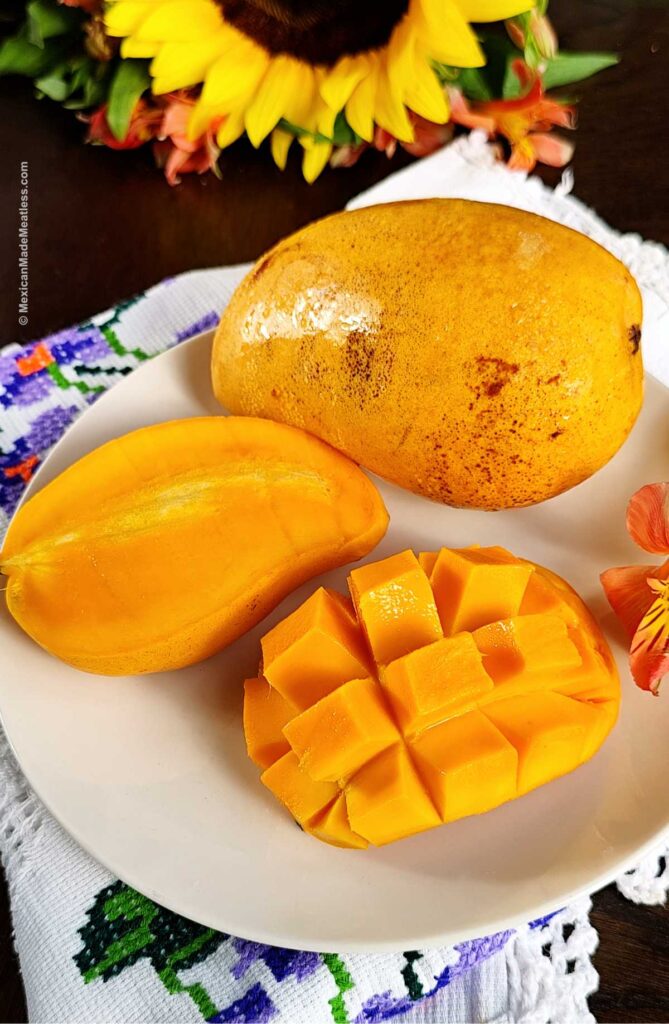 One whole Ataúlfo mango and one sliced in half and another piece cubed still inside the peel, all on a white plate.