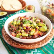Small white bowl filled with bean and avocado salad, there's a small brown bowl filled with corn chips and tostadas, two avocado halves, one large glass mixing bowl filled with the salad all on top of white kitchen countertop.
