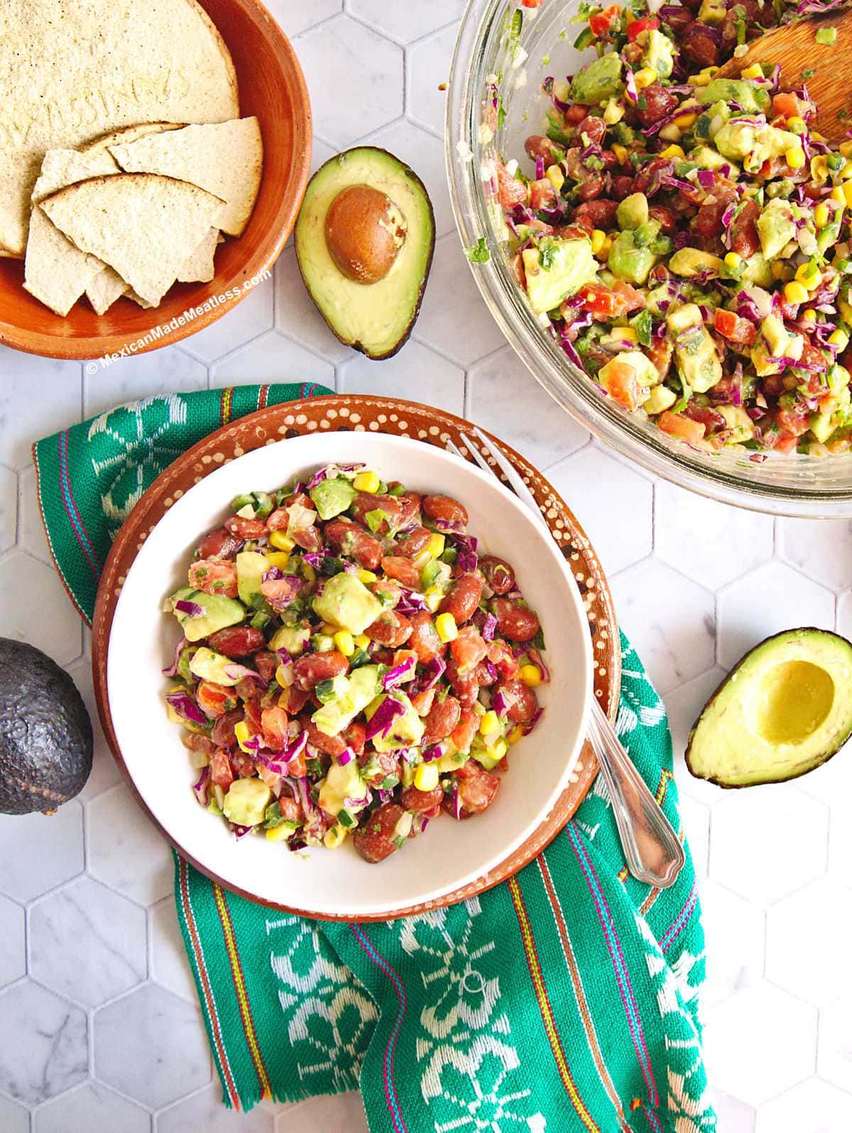 Small white bowl filled with bean and avocado salad, there's a small brown bowl filled with corn chips and tostadas, two avocado halves, one large glass mixing bowl filled with the salad all on top of white kitchen countertop.