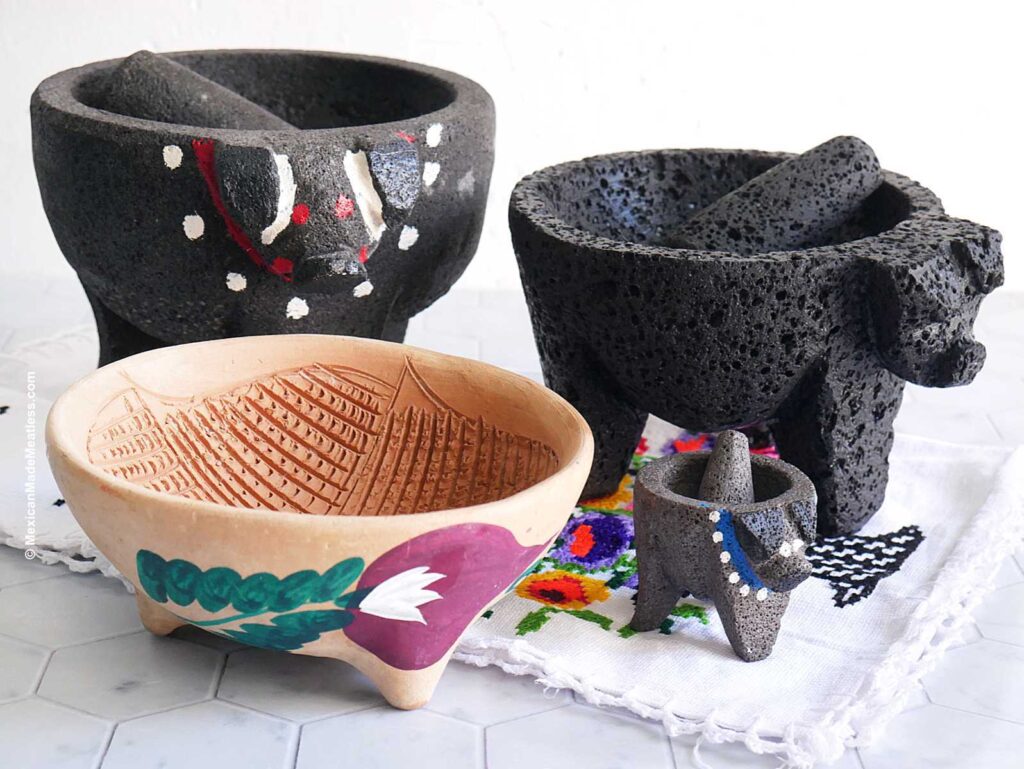 Three types of Mexican molcajetes on a white table. There is one made from lava rock, two made from stone and the other is a hand painted barro or terracotta molcajete.