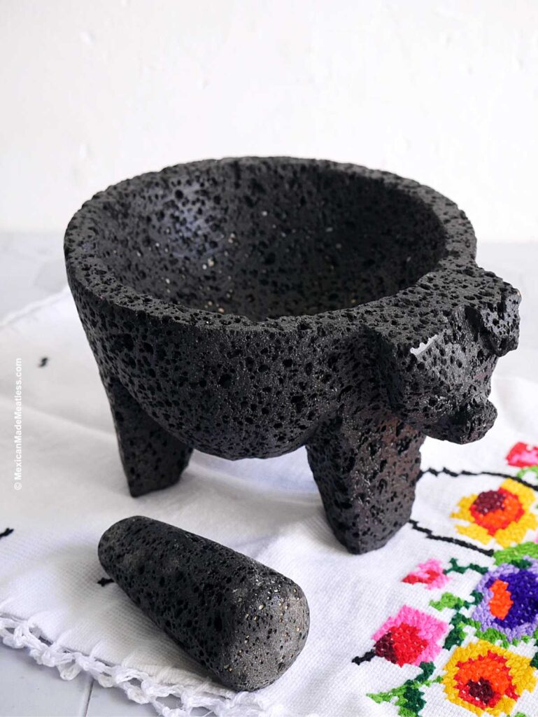 A Mexican molcajete made of lava rock with the pestle and both laying on top a decorative napkin.